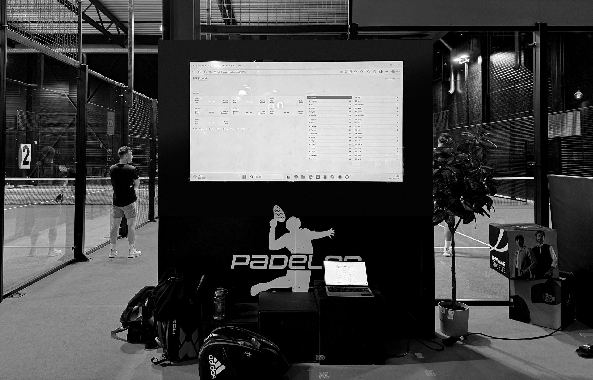 A big screen in an indoor padel hall showing the courts of the current round and the live standings